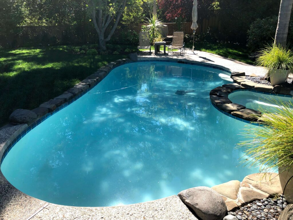 A kidney-shaped backyard pool is surrounded by rocks and trees, with two lounge chairs and a table on a sunny day.