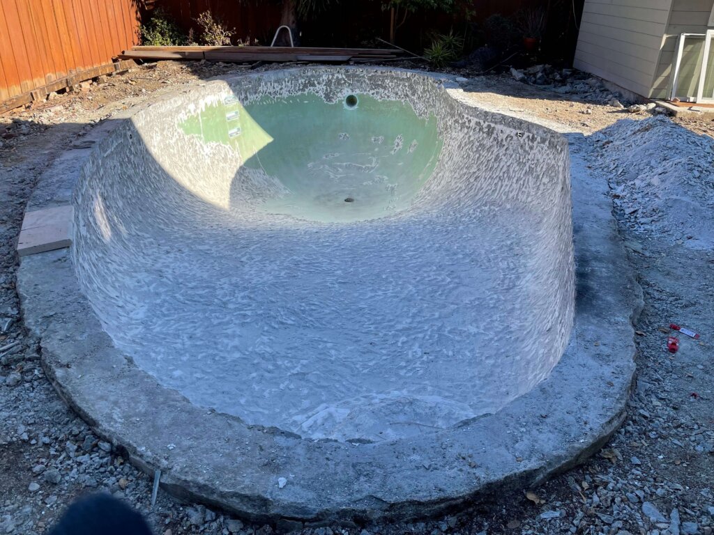 An empty, unfinished swimming pool with exposed concrete sits in a backyard under daylight. Construction materials are scattered around the area.