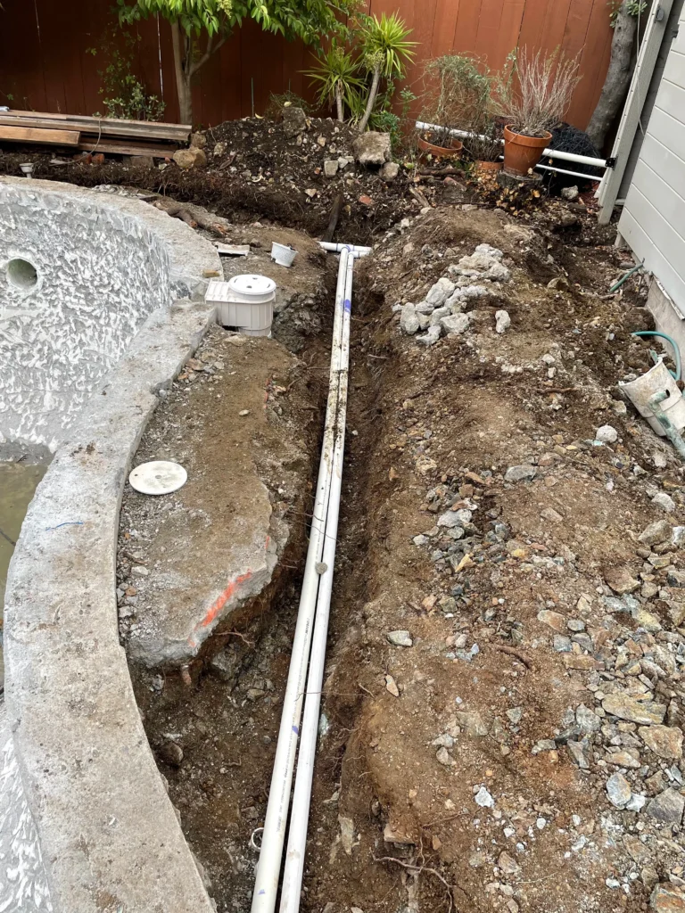 A trench with PVC pipes runs alongside an empty, partially demolished pool surrounded by dirt, rocks, and some garden plants.