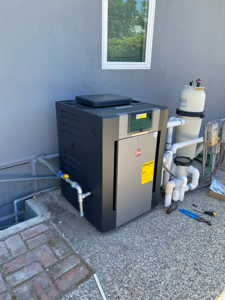 Outdoor tankless water heater installed next to a building wall, surrounded by plumbing pipes and pool equipment, with tools and a cardboard box nearby.