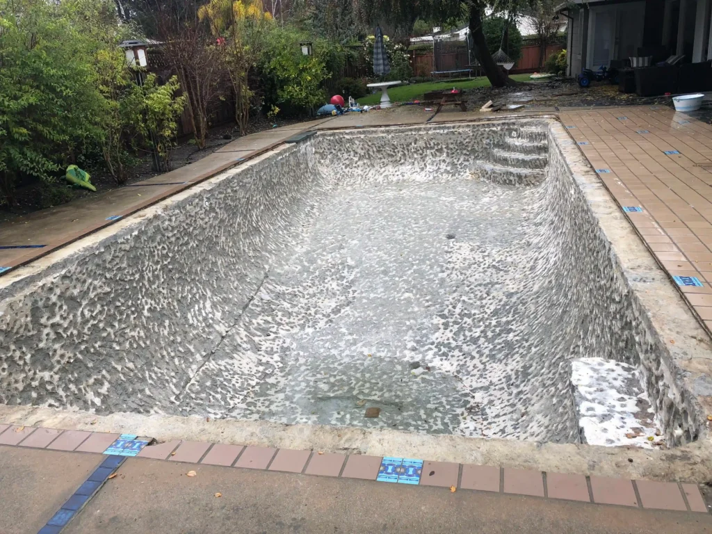 Empty, unfinished in-ground swimming pool with a rough, patchy surface, surrounded by a tiled deck. Trees and garden items are visible in the background.